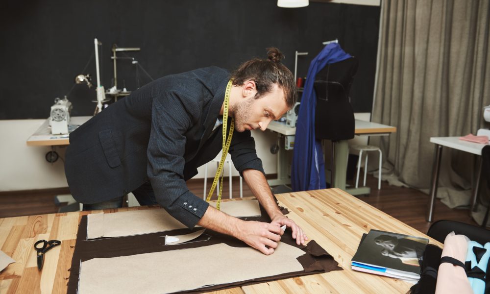 Portrait of serious young good-looking hispanic fashion designer in black suit cutting out parts of jacket for winter collection with concentrated face expression